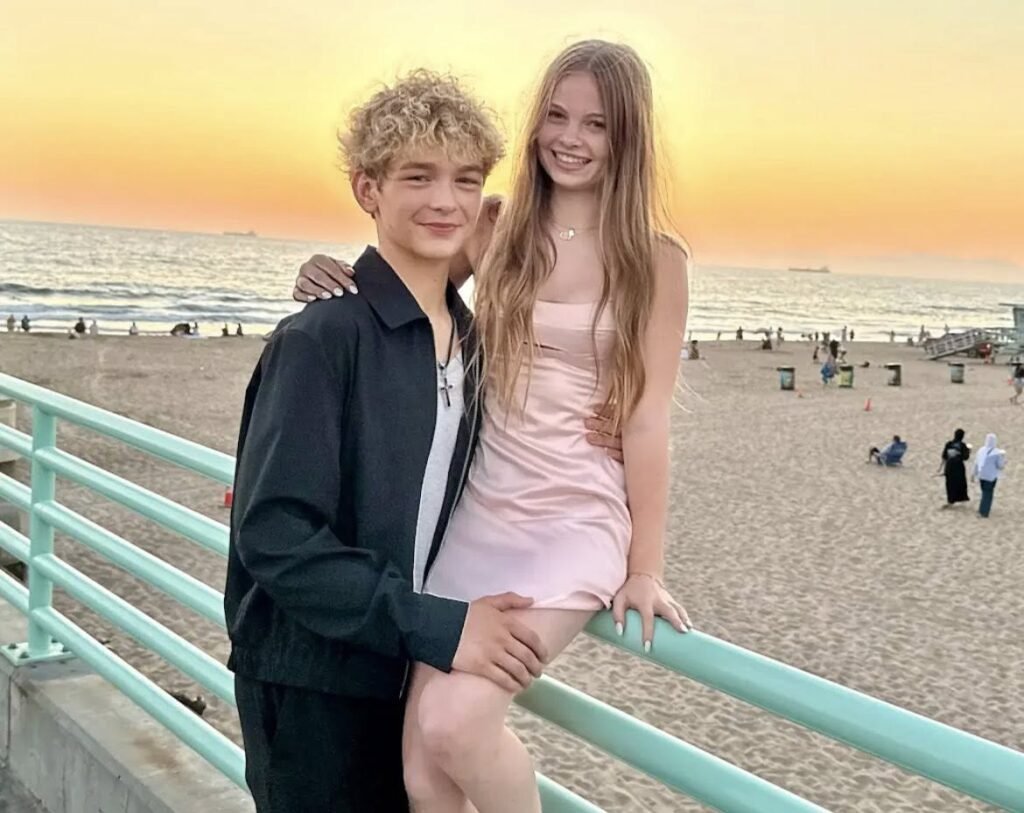 A candid, warm-toned photograph of teenage influencers Kaido and Salish posing together on a pier railing at sunset. Kaido, with curly blonde hair, wears a dark jacket and smiles slightly while embracing Salish. Salish, with long blonde hair, sits on the railing in a light pink dress, smiling broadly at the camera. The background shows a crowded beach, the ocean with distant ships, and a vibrant orange-yellow sunset sky. This image captures the young couple enjoying a beautiful evening by the sea.